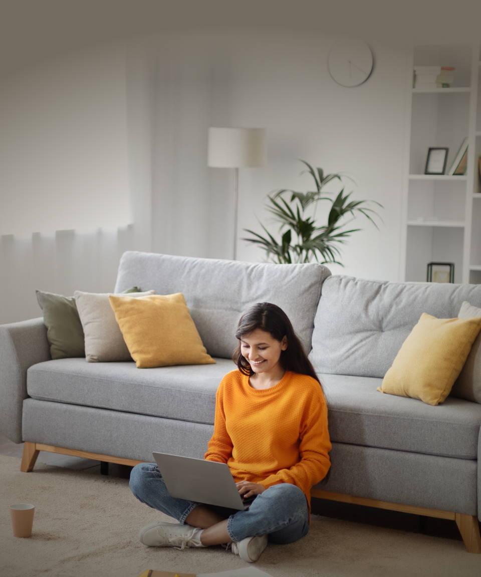 Woman sitting on the floor in front of her couch, cross-legged, smiling, typing on a laptop 