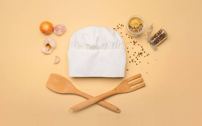 A chefs hat, wooden spatula and fork, onions and garlic, and spilled spices and peppercorns for Confetti's Cooking Classes for Beginners