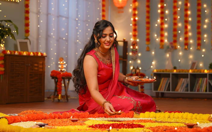 Indian woman sitting on the floor celebrating the holiday of Diwali, surrounded by bright colors, lights, and flowers.
