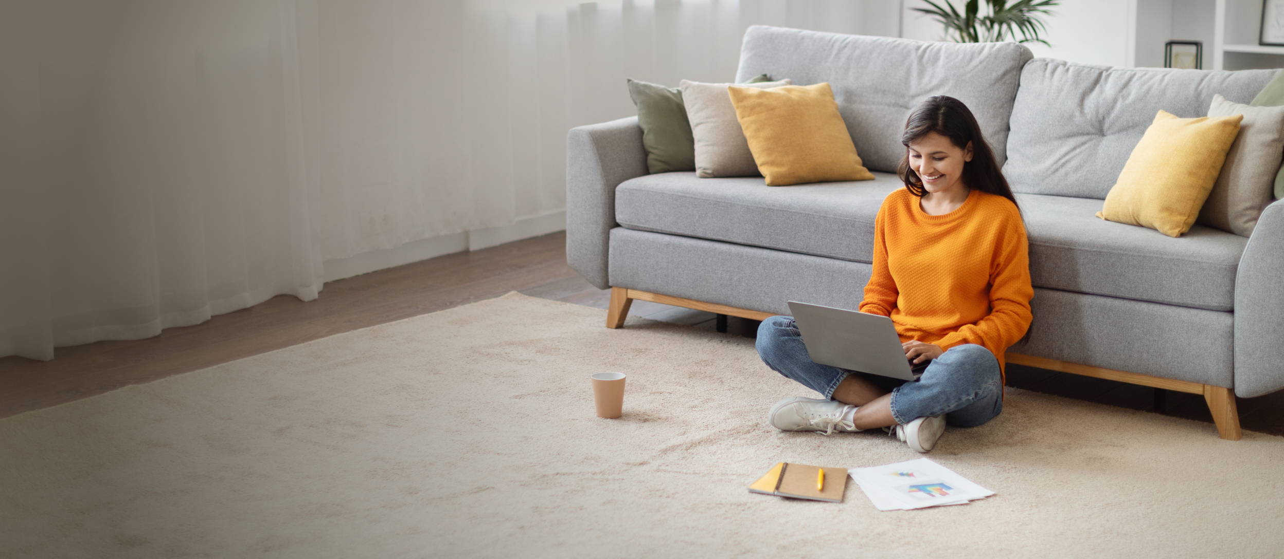 Woman sitting on the floor in front of her couch, cross-legged, smiling, typing on a laptop 