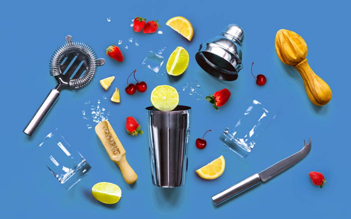 A cocktail shaker surrounded by various fruits, citrus reamers, glasses, ice, and a knife for Confetti's Virtual Mixology Classes