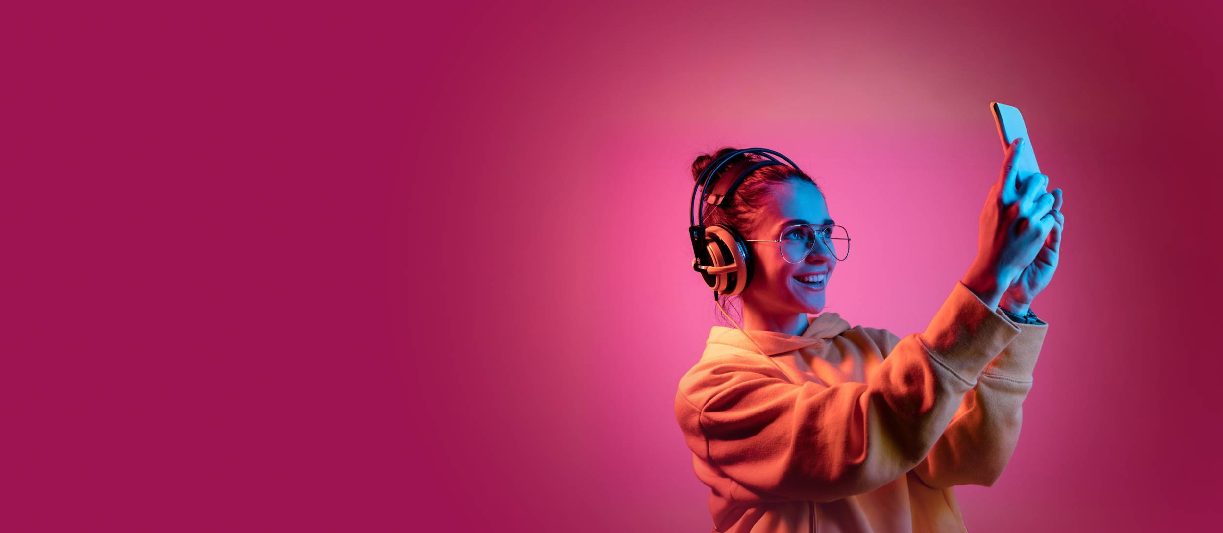 Smiling woman wearing headphones & an orange sweater, over a pink background, she's taking a photo