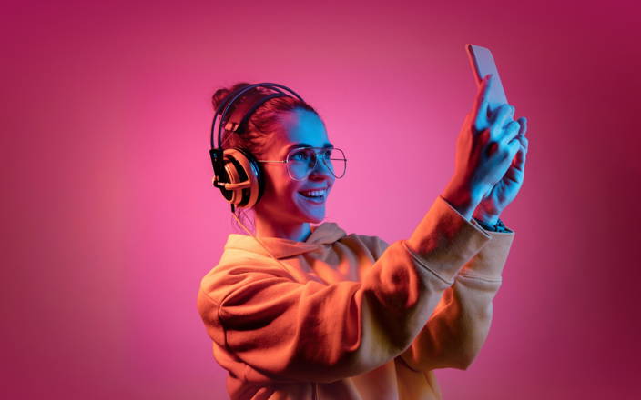 Smiling woman wearing headphones & an orange sweater, over a pink background, she's taking a photo