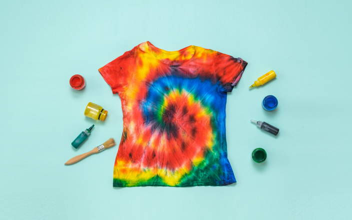 A tie dyed t-shirt surrounded by colorful ink dropper bottles and a paint brush for Confetti's Virtual Tie Dye Class