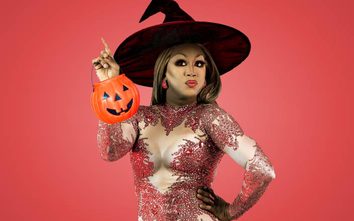 A drag queen wearing a witches hat and holding a jack-o-lantern bucket for Confetti's Virtual Halloween Drag Queen Trivia
