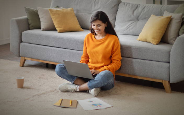Woman sitting on the floor in front of her couch, cross-legged, smiling, typing on a laptop 
