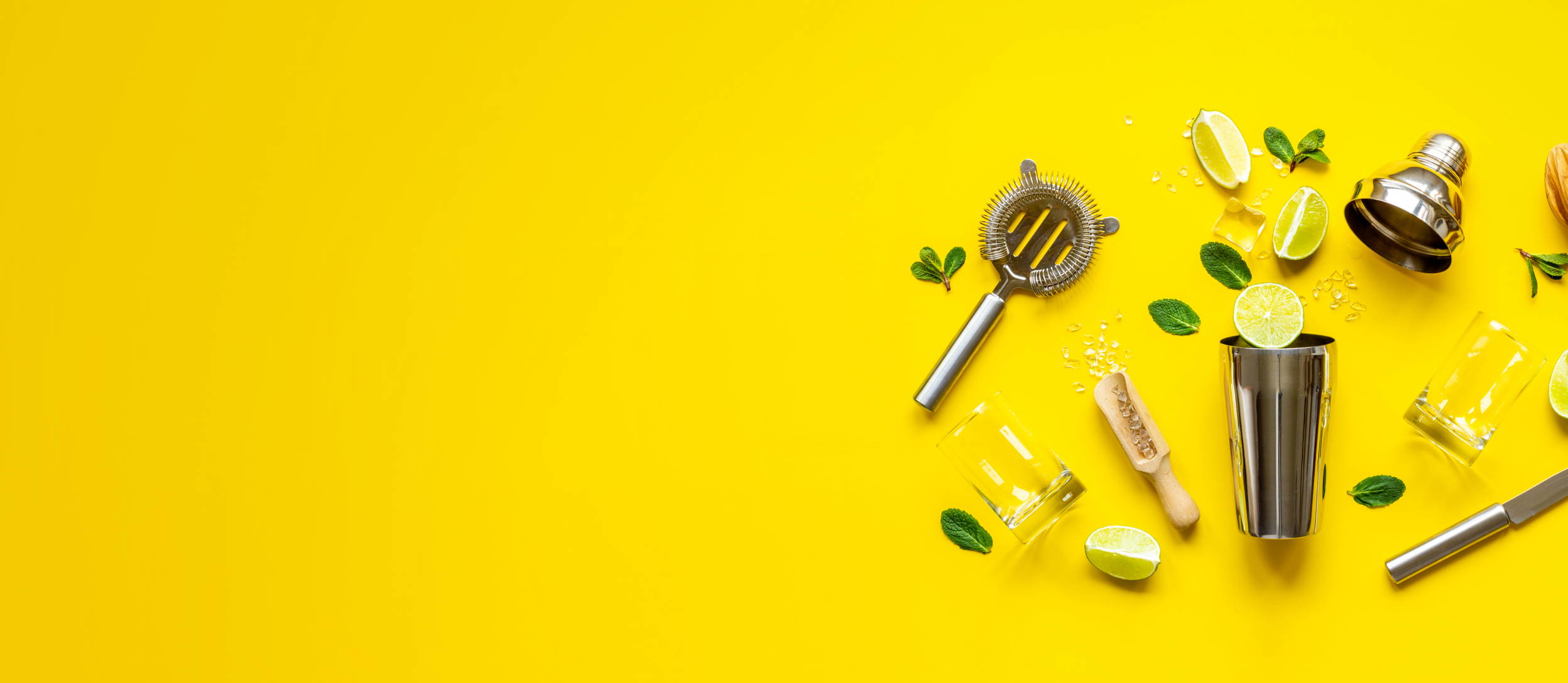 A cocktail shaker, strainer, knife, ice, a rocks glass, a scooper, sprigs of herbs, and slices of limes, all spread out for Confetti's BYO Mixology Class Team Building