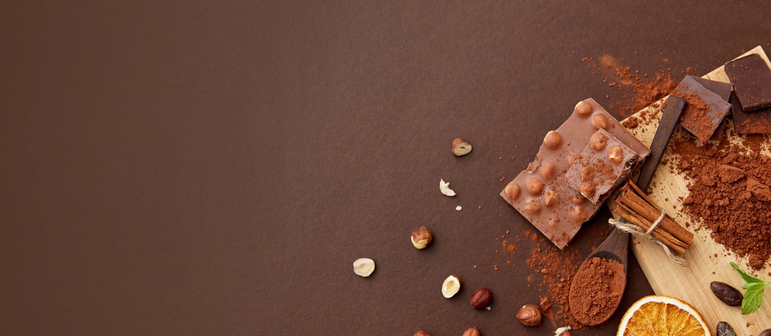 A cutting board with cocoa powder, cinnamon sticks, crushed hazelnuts, and a chocolate bar on it and scattered around for Confetti's Virtual Chocolate Tasting Team Building